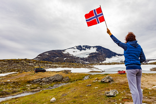Woman holds norwegian flag in mountains, van with canoe on top roof in distance. National tourist scenic route 55 Sognefjellet, Norway