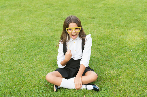 Small schoolgirl sit on green grass. Fashion accessory. Fashion style. Rock this year. Happy girl hold prop glasses. Beauty look of fashion model. Little child school uniform. Back to school.