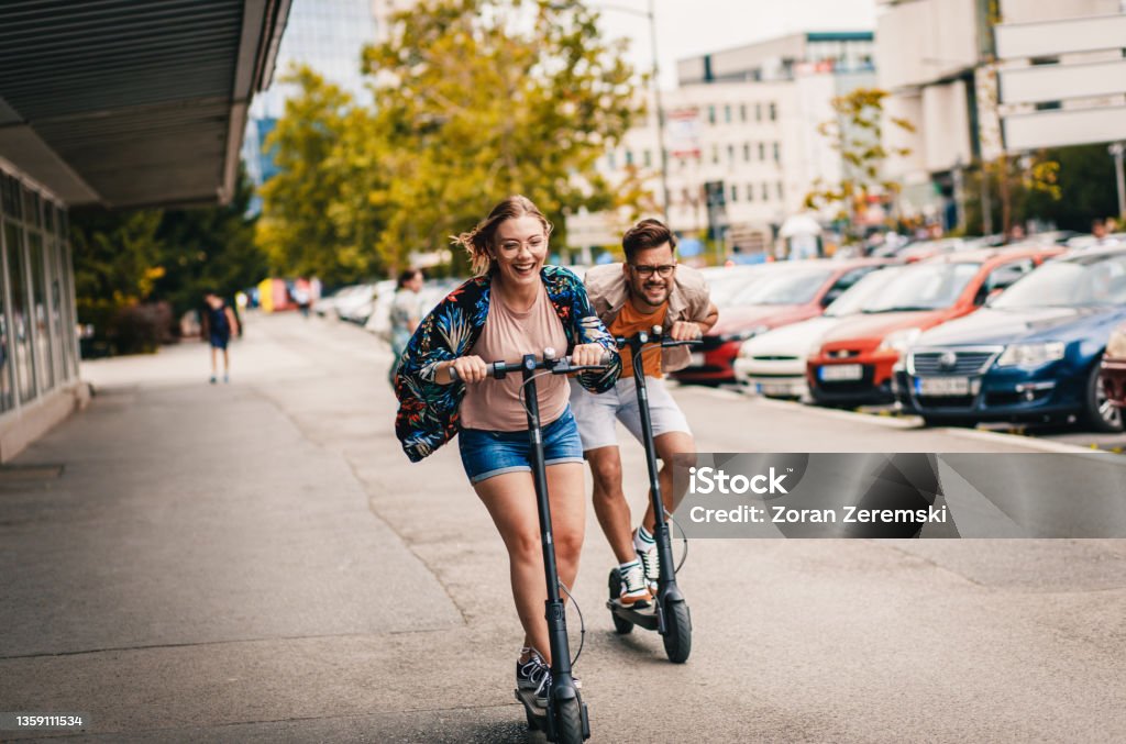 Young couple on vacation having fun driving electric scooter through the city. Electric Push Scooter Stock Photo