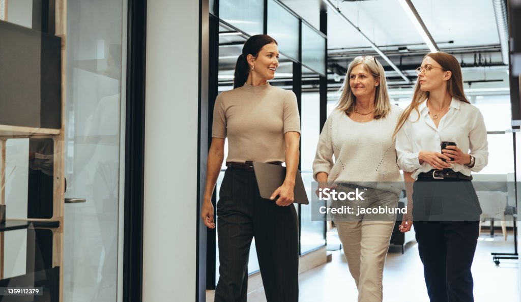 Three businesswomen walking together in an office Three cheerful businesswomen walking together in an office. Diverse group of businesswomen smiling while having a discussion. Successful female colleagues collaborating on a new project. Women Stock Photo