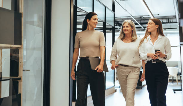 Three businesswomen walking together in an office Three cheerful businesswomen walking together in an office. Diverse group of businesswomen smiling while having a discussion. Successful female colleagues collaborating on a new project. three people stock pictures, royalty-free photos & images