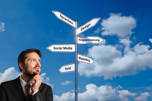 businessman standing in front of a signpost with many possible professions in German like ARTS, MARKETING, SOCIAL MEDIA, PROGRAMMING, DESIGN, LANGUAGES with a cloudy background