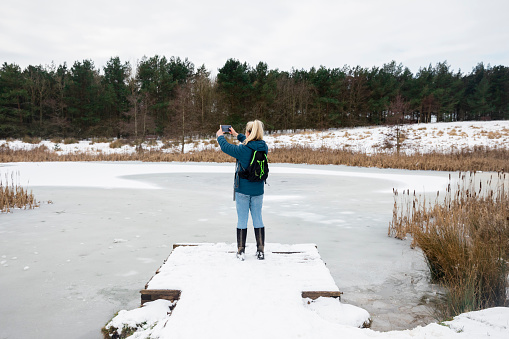 A wide angle rear view of a blonde female stood at the end of a jetty in a nature reserve in Cramlington in the north-east of England. She is stood taking a picture on her mobile phone of the beautiful landscape and frozen lake.