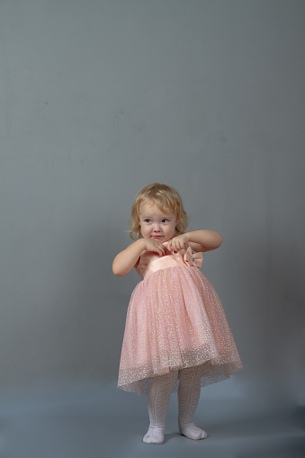 Cute baby girl 2 years old caucasian dancing in pink prom dress on a gray background in the studio.
