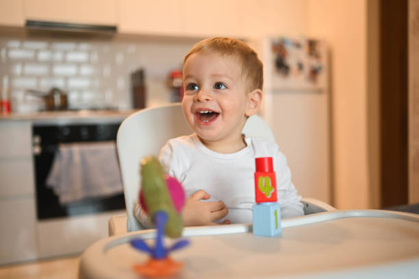 Little happy cute baby toddler boy blonde sitting on baby chair playing with constructor. Baby facial expressions indoors at home kitchen interior with toys. Healthy happy family childhood concept. stock photo