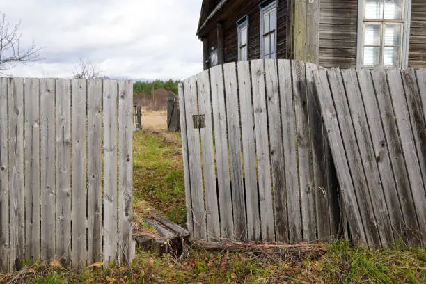 Photo of Broken rickety wooden fence near old house in Russian village. Autumn. Grey sky