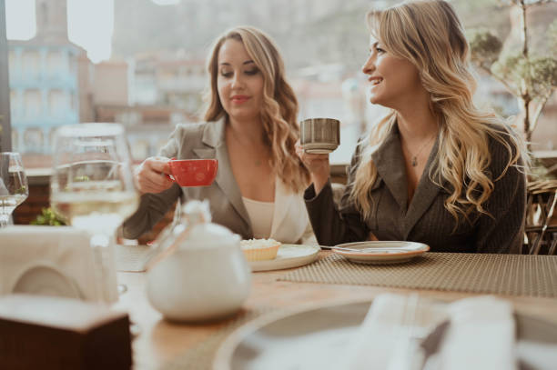 Close-up portrait of two female friends in strict suits laughing drinking coffee and wine on the terrace outside at summer street cafe on background buildings of Old Tbilisi city, Georgia stock photo