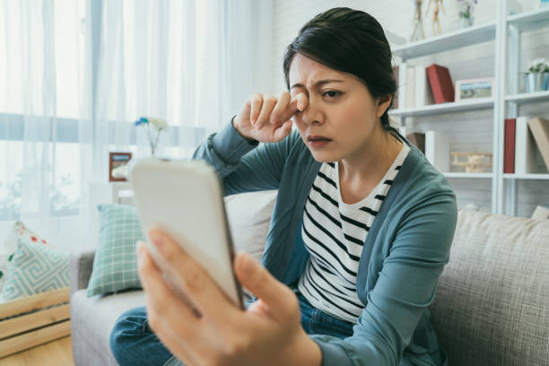 la donna asiatica con messa a fuoco selettiva che soffre di visione offuscata si strofina gli occhi stanchi mentre tiene il suo smartphone sul divano nel soggiorno di casa. - strizzare gli occhi foto e immagini stock