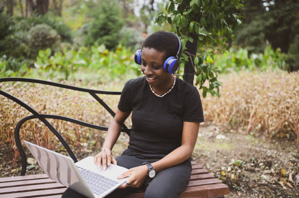 Smiling happy African black short-haired woman student afro hair with blue headphones studying online working on laptop computer at summer green park. Diversity. Remote work, distance education. stock photo