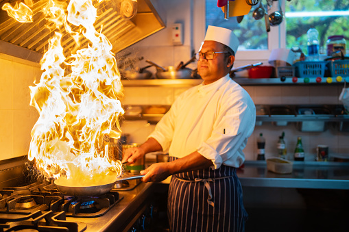A chef watching the flames in the frying pan as he flambés the food he is cooking at the stove in a restaurant kitchen