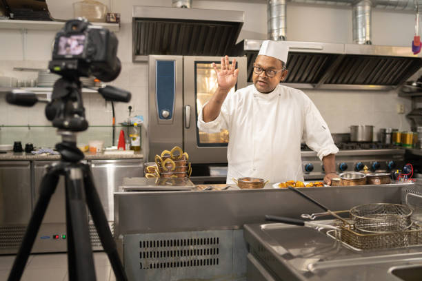 A professional chef waving to the camera During an online cookery virtual event, the professional Indian chef waves to the camera set up in front of him in a large commercial kitchen caterer photos stock pictures, royalty-free photos & images