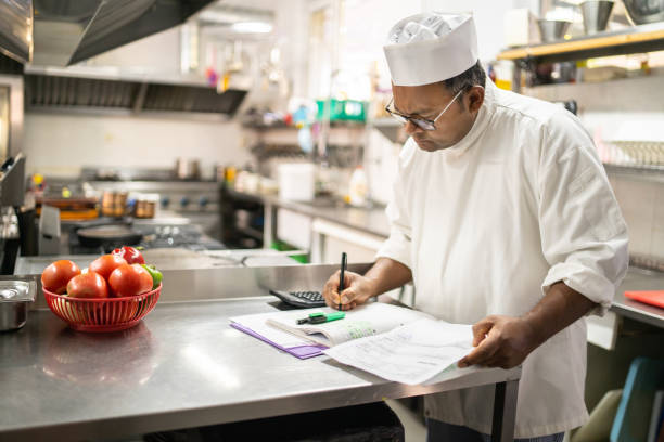 A chef is writing in the account book Using a stainless steel kitchen counter, a chef is writing the numbers into an accounting book for the kitchen finances demanding photos stock pictures, royalty-free photos & images