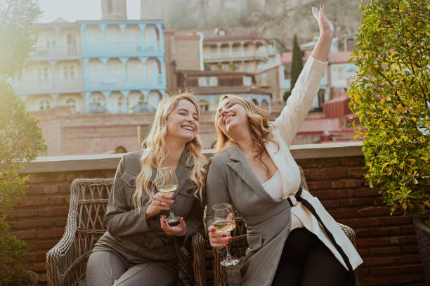 Close-up portrait of two female friends in strict suits laughing drinking wine on the terrace outside at summer street cafe on background buildings of Old Tbilisi city, Georgia. stock photo