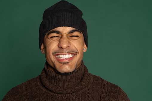 Laughing. Close-up portrait of African young man in warm clothes and hat isolated on green color studio background. Concept of emotions, expression, youth, sales, diversity. Copy space for ad