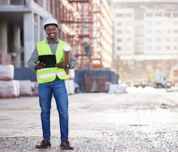 retrato completo de um belo jovem trabalhador da construção civil em pé com plantas e prancheta em um canteiro de obras - full length clipboard african ethnicity black - fotografias e filmes do acervo