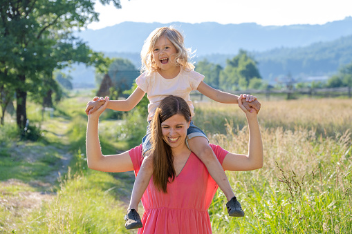 Woman carrying her daughter on shoulders. Smiling females enjoying time outdoors together. Golden hour in summer nature.