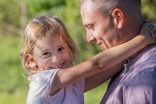 Caucasian school age blonde girl holding dad around neck. Looking at camera and smiling. Romantic close-up.