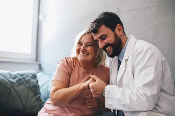A young bearded doctor is sitting on the sofa, hugging a senior lady and holding her hand. Both of them are smiling. A senior lady is happy because she was concerned about her health, but when the doctor finished the examination, she was released, because all results were fine.