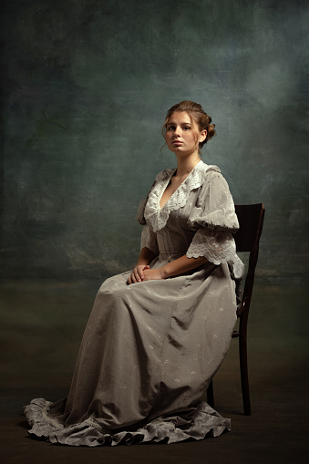 Sad young beautiful girl in gray dress of medieval style sitting on chair isolated on dark vintage background. Comparison of eras concept, flemish style. Classic art character, old-fashioned.