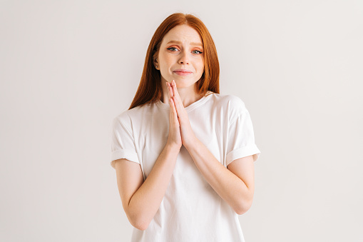 Portrait of crying young woman makes imploring expression keeps palms pressed together asks for favor says please looking at camera with hope, standing on white isolated background in studio.