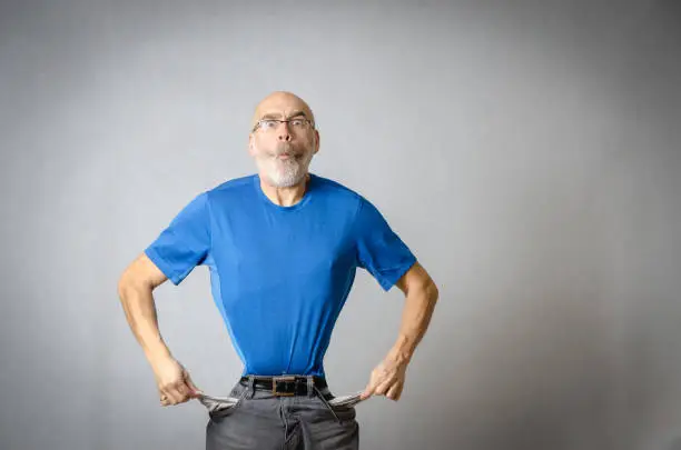 Mature man showing his empty pocket, his tighten belt and waist and hollow cheeks to express he is lacking money and has difficult financial times