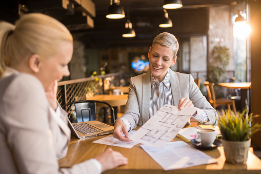 Happy real estate agent showing her customer an apartment blueprint in a cafe.
