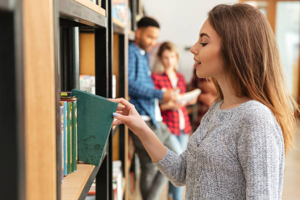 istock Young pretty woman student standing in library holding book. 1359194548