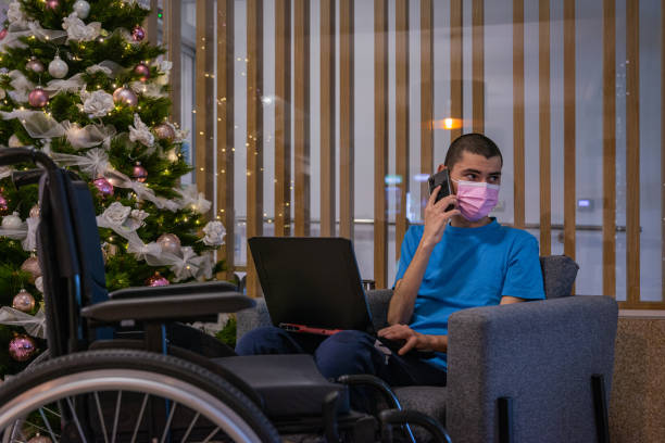 A teenager with a disability — with dystrophy in a blue T-shirt in a wheelchair with a medical mask due to covid next to a richly decorated and lit Christmas tree in the hotel lobby — works on a computer, makes a phone call, and looks at a Christmas tree. un adolescente con una discapacidad, con distrofia en una camiseta azul en una silla de ruedas con una máscara médica debido a covid junto a un árbol de navidad ricamente decorado e iluminado en el vestíbulo del hotel, trabaja en una computadora, hace