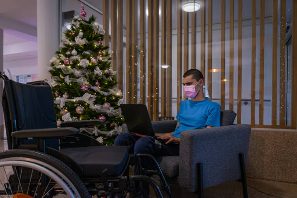 A teenager with a disability — with dystrophy in a blue T-shirt in a wheelchair with a medical mask due to covid next to a richly decorated and lit Christmas tree in the hotel lobby — works on a computer, makes a phone call, and looks at a Christmas tree. un adolescente con una discapacidad, con distrofia en una camiseta azul en una silla de ruedas con una máscara médica debido a covid junto a un árbol de navidad ricamente decorado e iluminado en el vestíbulo del hotel, trabaja en una computadora, hace