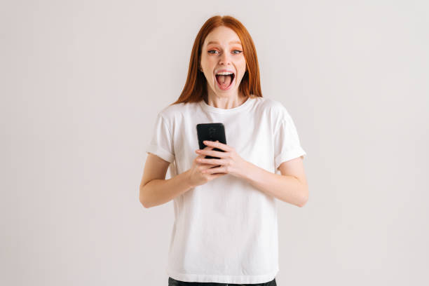 retrato de estudio de una joven alegre con la boca abierta leyendo un buen mensaje en línea usando el teléfono móvil sobre un fondo blanco aislado. - regocijo fotografías e imágenes de stock