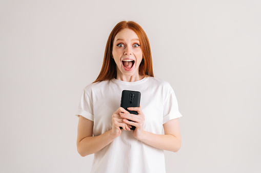 Studio portrait of happy excited young woman open mouth reading good online message using mobile phone on white isolated background. Cheerful lady holding smartphone in hands and looking at camera.