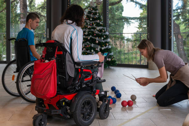Two disabled people in wheelchairs play boccia next to a decorated Christmas tree with a beautiful view through the windows of the terrace of the green trees in the evil seaside resort.