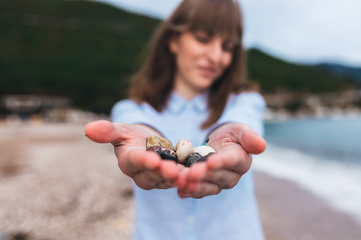 A young brunette woman holding seashells in her hands at the beach.