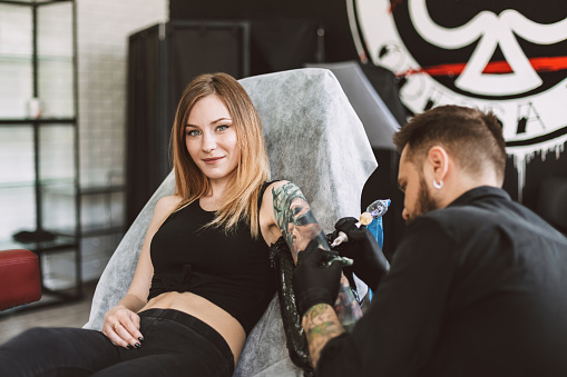 Smiling tattooed girl looking in camera while professional tattooer doing tattoo on hand using tattoo machine in studio