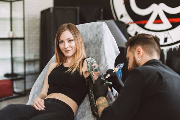 uśmiechnięta wytatuowana dziewczyna patrząca w kamerę profesjonalny tatuator robiący tatuaż na rękę za pomocą maszyny do tatuażu w studio - tattoo sleeve zdjęcia i obrazy z banku zdjęć