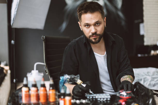 Professional tattooer in black gloves thoughtfully looking in camera holding tattoo machine in hand in modern studio Professional tattooer in black gloves thoughtfully looking in camera holding tattoo machine in hand in modern tattoo studio arm tattoo ideas for men pictures stock pictures, royalty-free photos & images