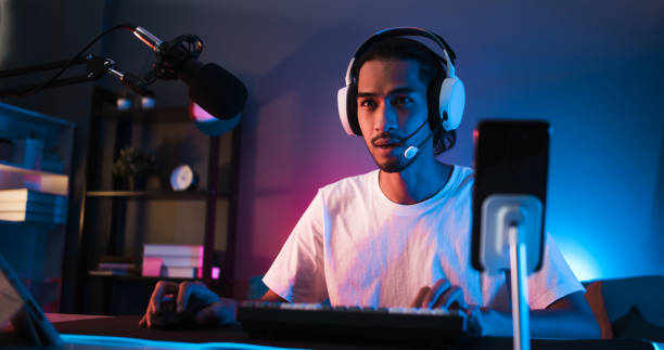 Young confident Asian man playing online computer video game, colorful lighting broadcast streaming live at home. Gamer lifestyle, E-Sport online gaming technology concept stock photo