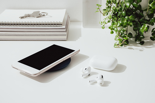 Charging mobile phone at home office desk with a keys, books, plant, bluetooth speaker and earphones.