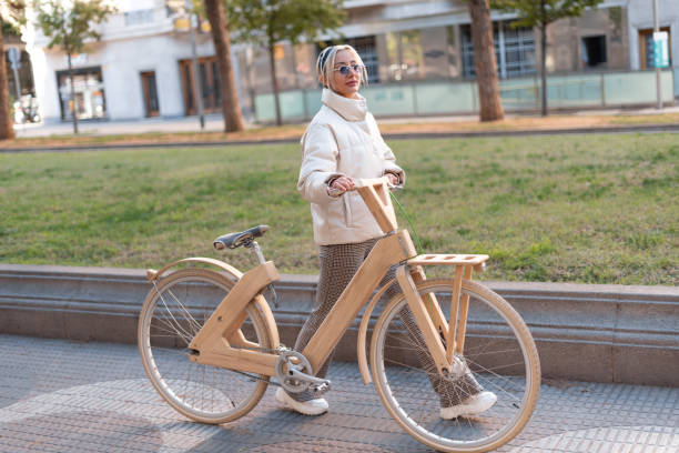 Full body blond woman in outerwear pushing wooden eco bike while walking on pavement near lawn in city park.