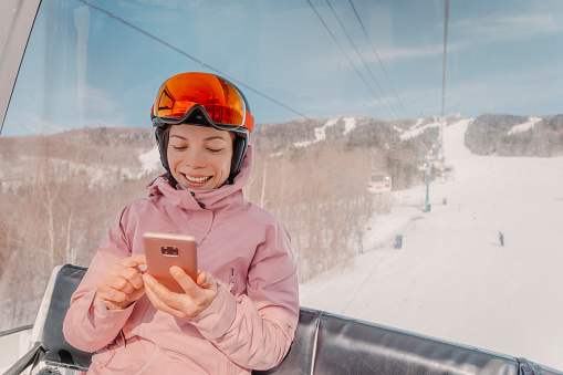 Ski holidays - Woman skier using phone app in gondola ski lift. Girl smiling looking at mobile smartphone wearing ski clothing, helmet and goggles. Ski winter activity vacation concept.