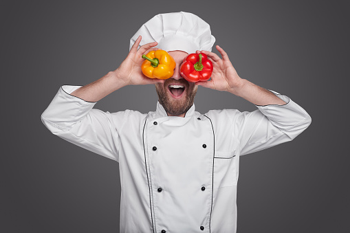 Funny bearded male chef in white uniform covering eyes with fresh yellow and red peppers and smiling brightly, while preparing healthy food against gray background