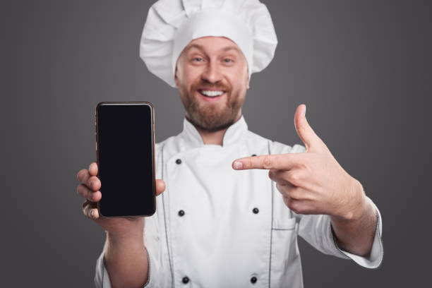 istock Smiling chef in uniform pointing at smartphone 1359248136