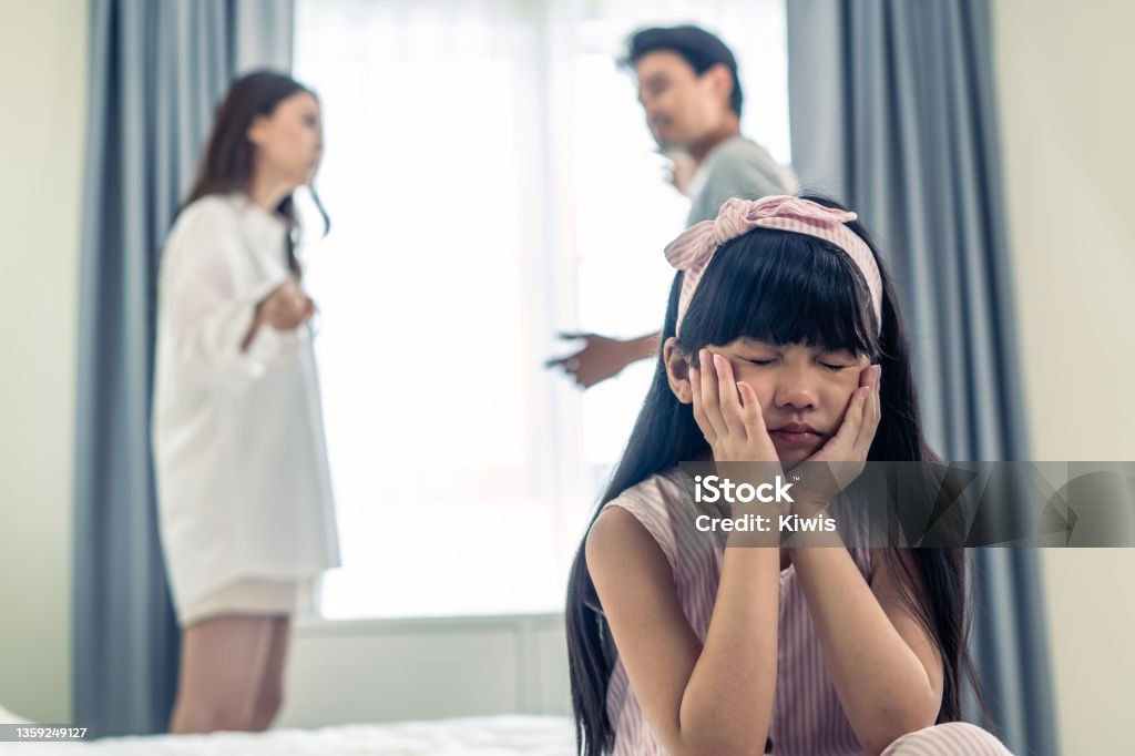 Asian little girl crying while parents fighting or quarrel conflict. Young kid daughter sit on bed feel heart broken with tears and dont want to see violence at home. Family problem-separation concept Anger Stock Photo