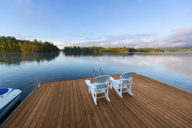 Beautiful morning landscape panorama of a lake in cottage country in Ontario, Canada Beautiful morning landscape panorama, under a blue sky, of a lake in cottage country in Ontario, Canada. Two white Adirondack chairs facing the calm water are sitting on a new wooden dock. cottage stock pictures, royalty-free photos & images
