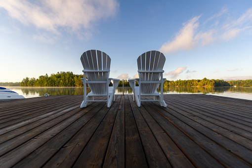 Scenic morning view of a bay in Jack's Lake,  Ontario Canada, from a cottage wooden dock. Two white Adirondack chairs are sitting on the pier facing the calm waters. Some clouds are visible in the sky.