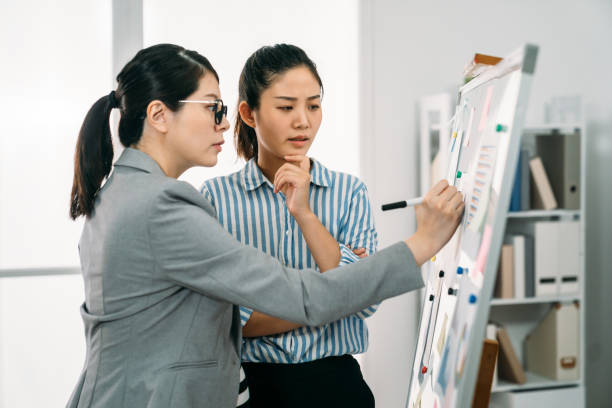 istock businesswoman is rubbing chin with thoughtful face while looking at the notes her expectant coworker is writing on whiteboard during discussion in the office. 1359264389