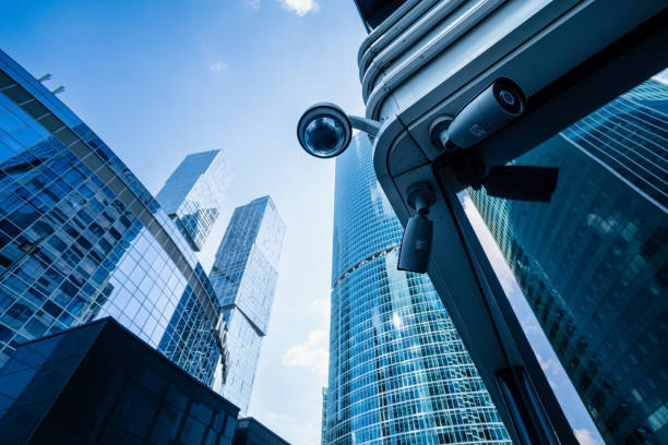 A low-angle view of modern skyscrapers with reflective glass facades under a blue sky, featuring a security camera mounted on a building in the foreground.