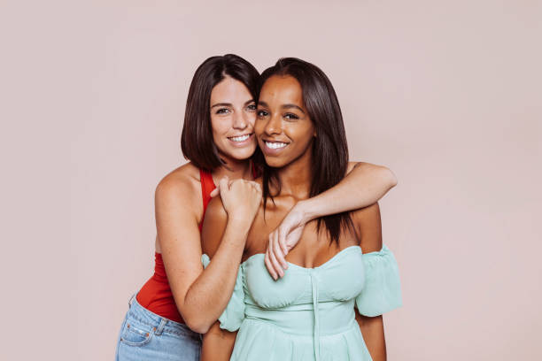 Two multiracial female friends hugging at studio Mixed race and caucasian woman hugging at studio. Two cheerful multiracial female friends posing and looking at camera over beige background. Happy lifestyle concept. isolated color stock pictures, royalty-free photos & images