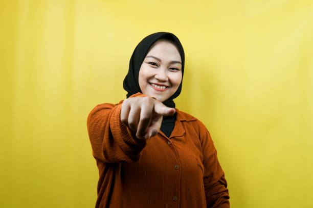 Beautiful young asian muslim woman with hands pointing at camera, wanting you, pointing at you, wanting to be with you, smiling confident, enthusiastic and cheerful, facing the camera, isolated, advertising concept stock photo