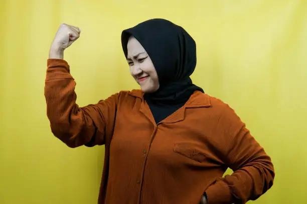 Photo of Beautiful asian young muslim woman with raised muscles, strength sign arms, isolated